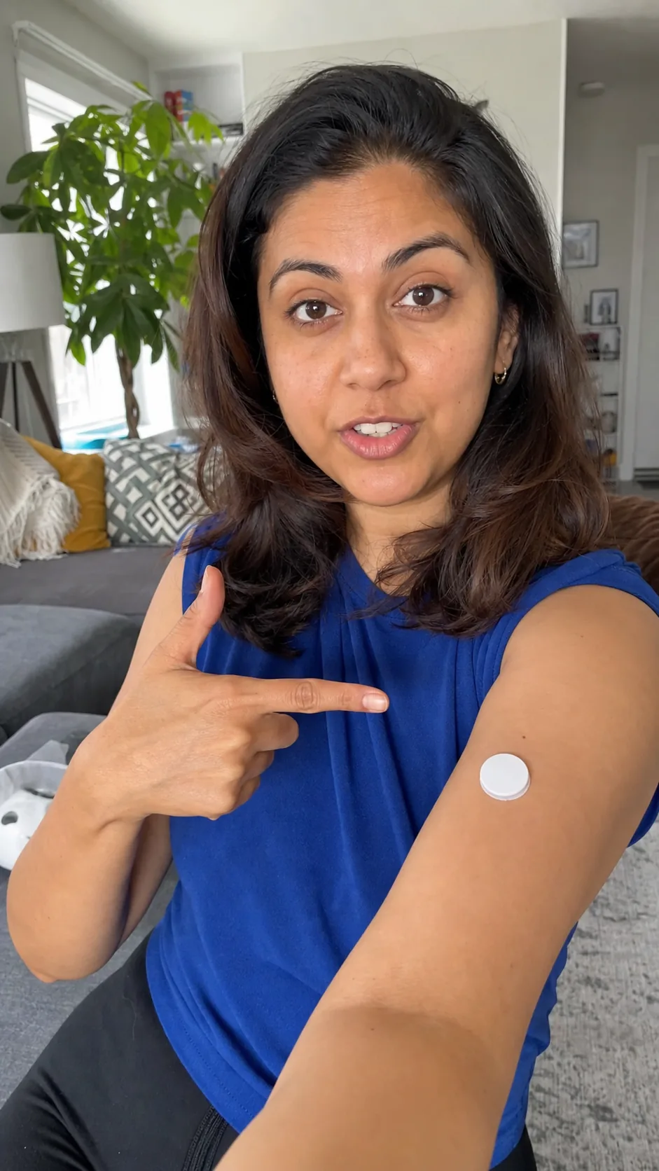 Woman in a living room points to a plain wearable sensor on her upper arm.